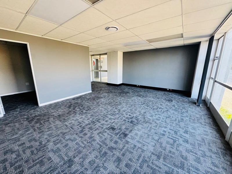A bright, open-plan office space featuring neutral grey walls, durable carpet tiles, a suspended ceiling, and large windows providing ample natural light.