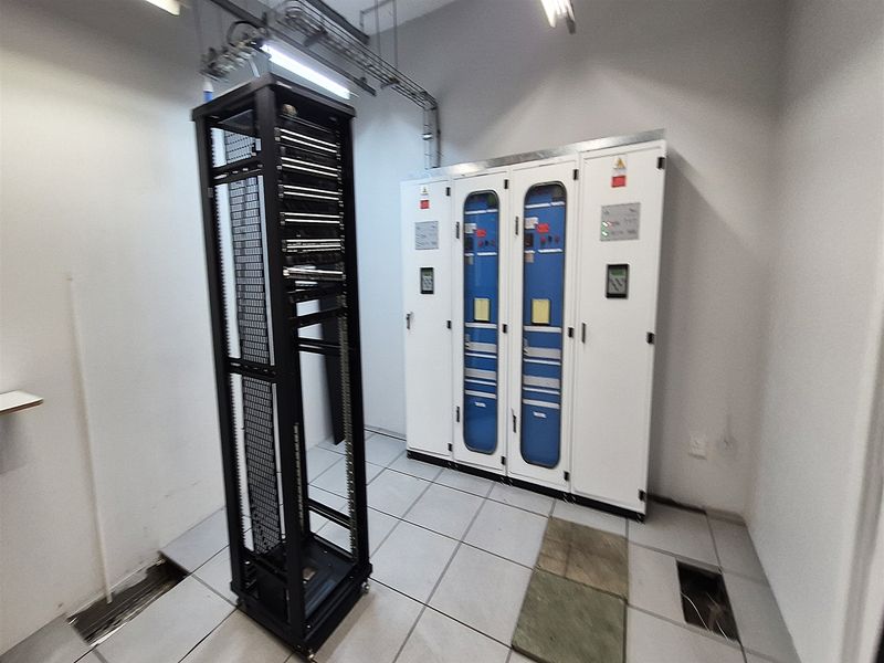 A functional utility or server room featuring industrial electrical distribution boards, a server rack, and a raised floor system with some maintenance access points.