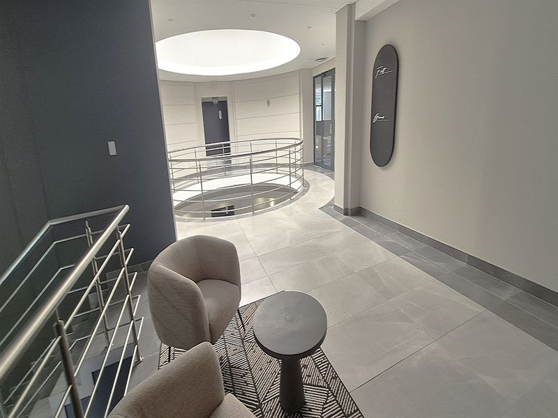 Modern office hallway featuring a circular atrium void, stainless steel balustrades, and high-quality tiled flooring with recessed ceiling lighting.