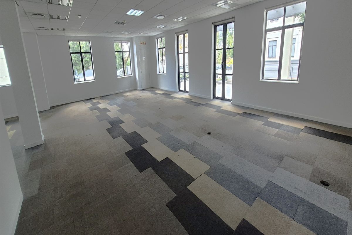 A spacious, empty office area featuring a suspended ceiling, recessed lighting, and modern patterned carpet tiles with ample natural light from large windows.