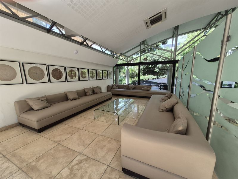 A modern office lounge area featuring tiled flooring, a sloped ceiling with integrated lighting, and large glass windows overlooking a garden.