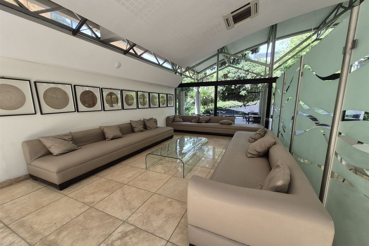 A modern office lounge area featuring tiled flooring, a sloped ceiling with integrated lighting, and large glass windows overlooking a garden.