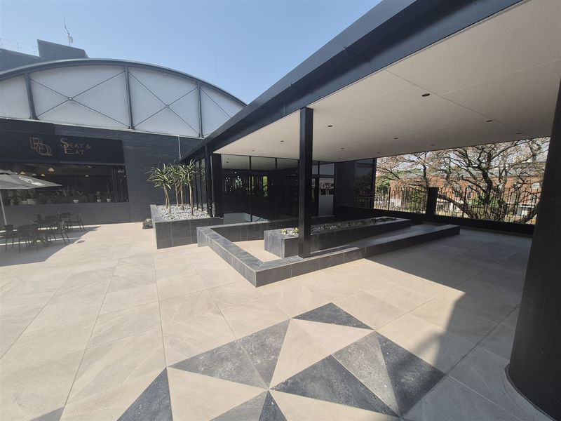 A modern, spacious covered outdoor patio area featuring geometric tiled flooring, built-in planters, and clean architectural lines, suitable for office breakout or social space.