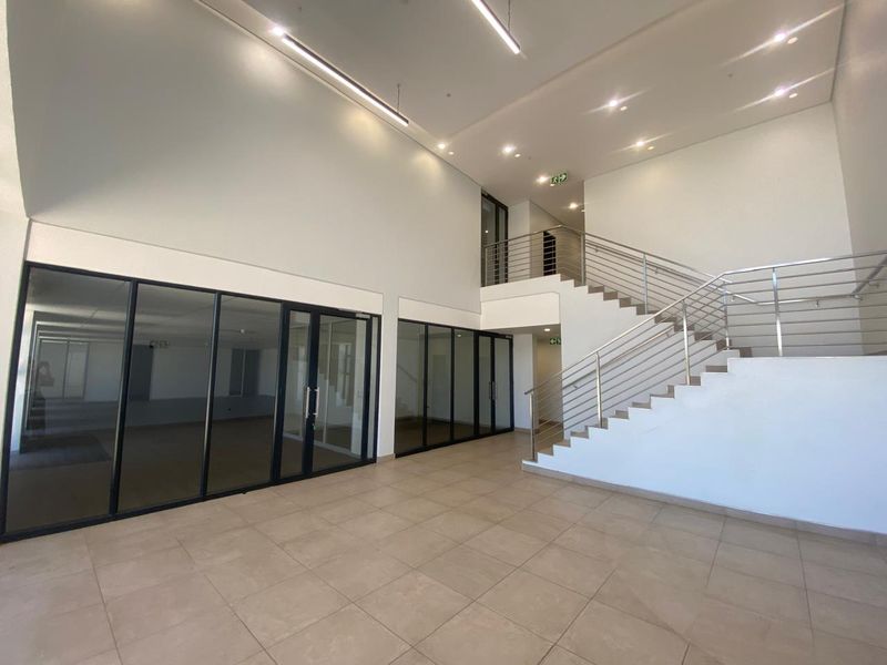 Spacious modern industrial entrance foyer featuring high ceilings, tiled flooring, a prominent staircase with stainless steel balustrades, and glass-partitioned office or meeting spaces.