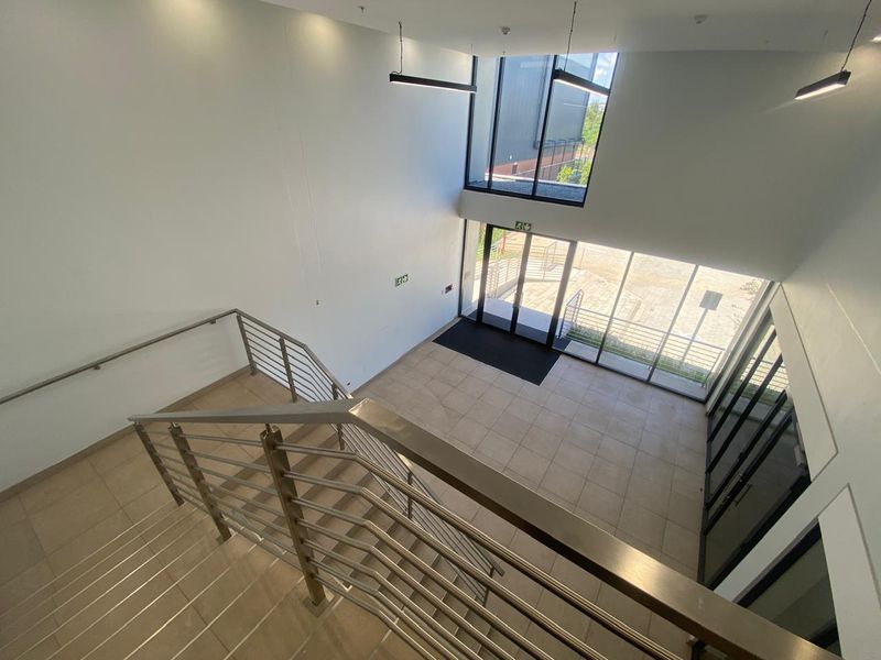 Modern double-volume industrial entrance foyer featuring tiled flooring, stainless steel staircase railings, and expansive glass windows providing abundant natural light.