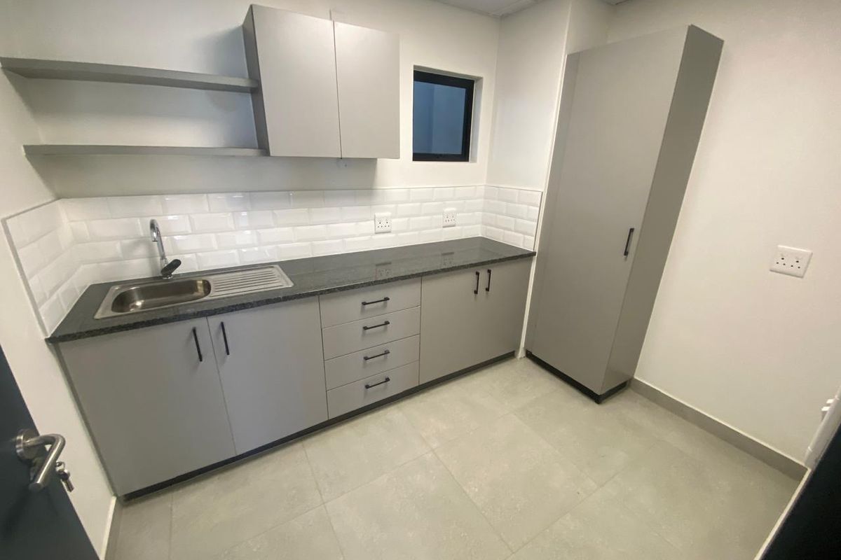 A clean, modern kitchenette featuring grey cabinetry, stone countertops, subway tile backsplash, and a stainless steel sink in an industrial office setting.