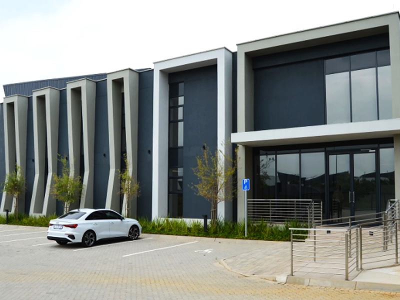 Modern industrial building facade featuring a glass entrance, architectural concrete fins, and a paved parking area with accessible ramp access.