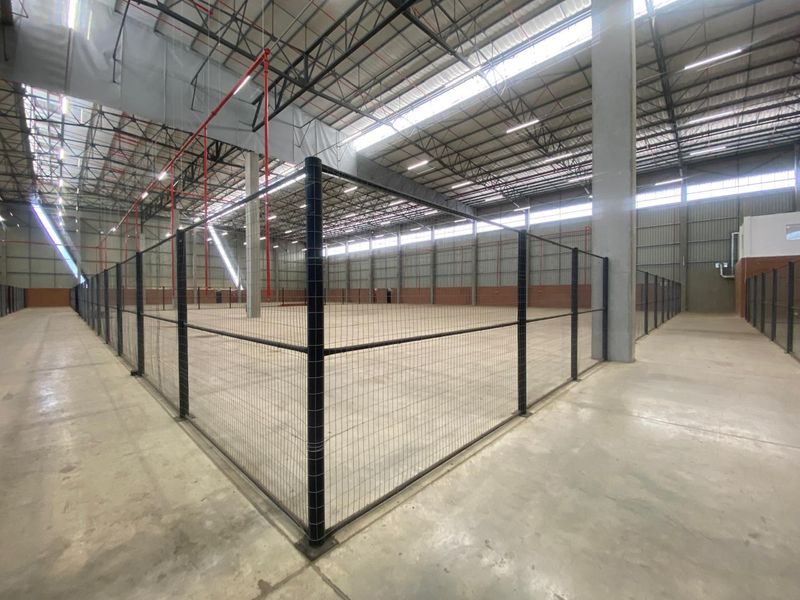 Spacious industrial warehouse interior featuring high ceilings with exposed steel trusses, concrete flooring, fire sprinkler system, and secure wire mesh partitioned areas.