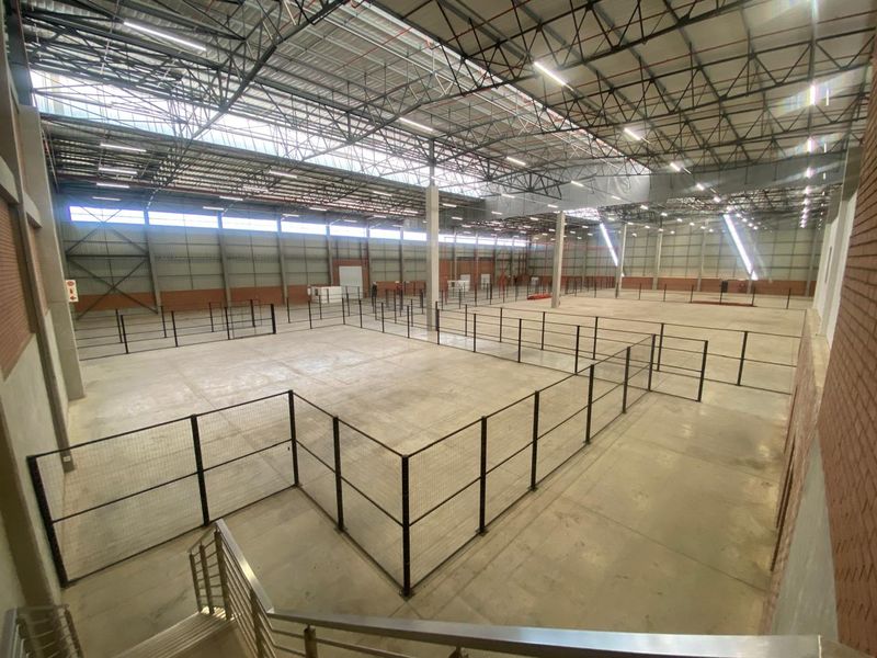 Expansive industrial warehouse space featuring high ceilings with exposed steel trusses, concrete flooring, and modular metal mesh partitioning throughout the open floor plan.