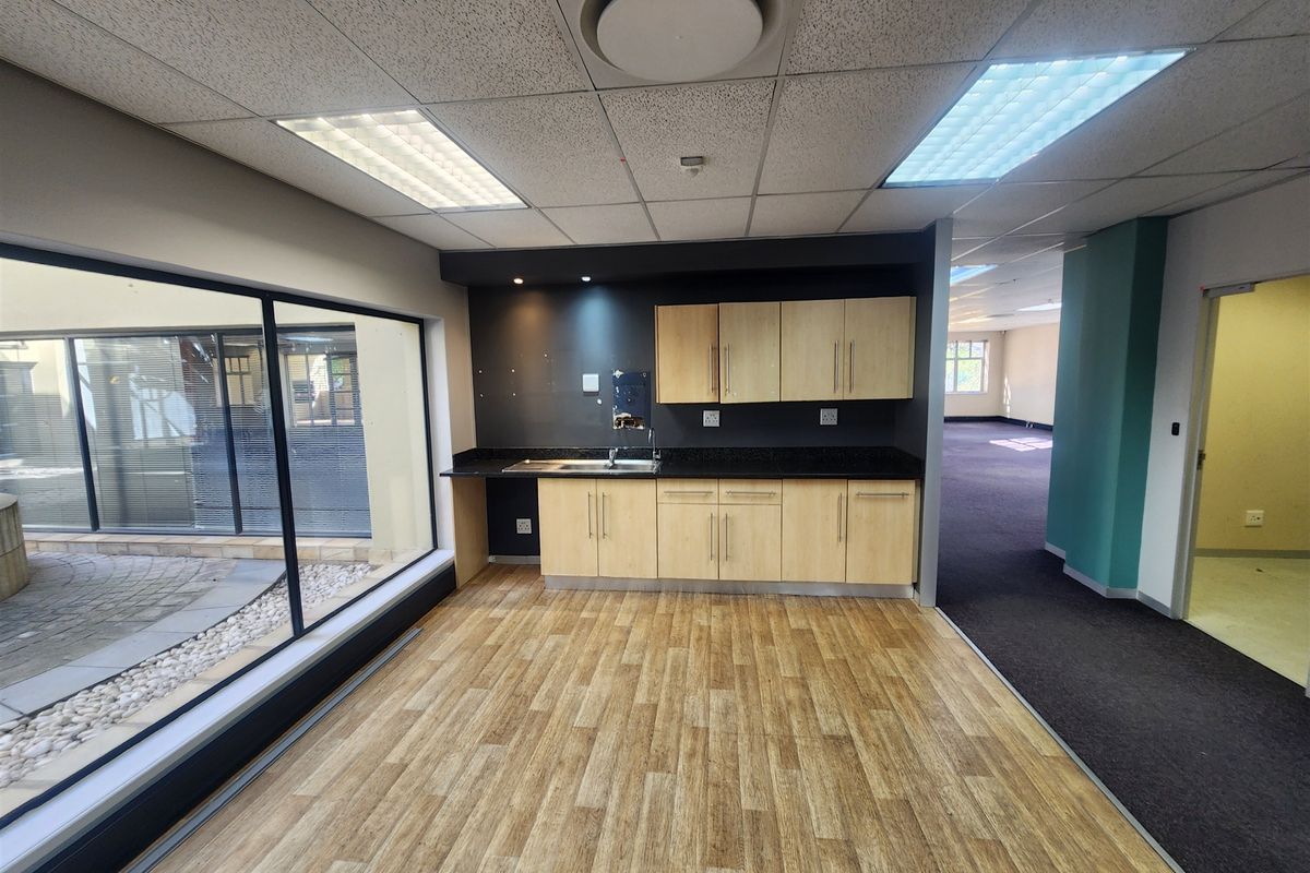 A clean, modern office kitchenette featuring light wood cabinetry, a dark countertop, and a large window overlooking an outdoor courtyard area.