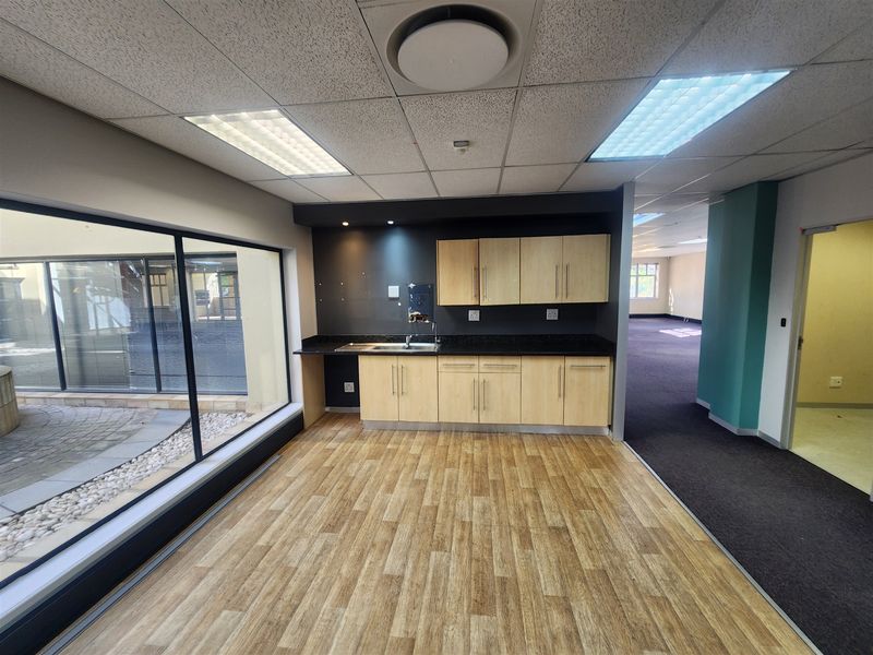 A clean, modern office kitchenette featuring light wood cabinetry, a dark countertop, and a large window overlooking an outdoor courtyard area.