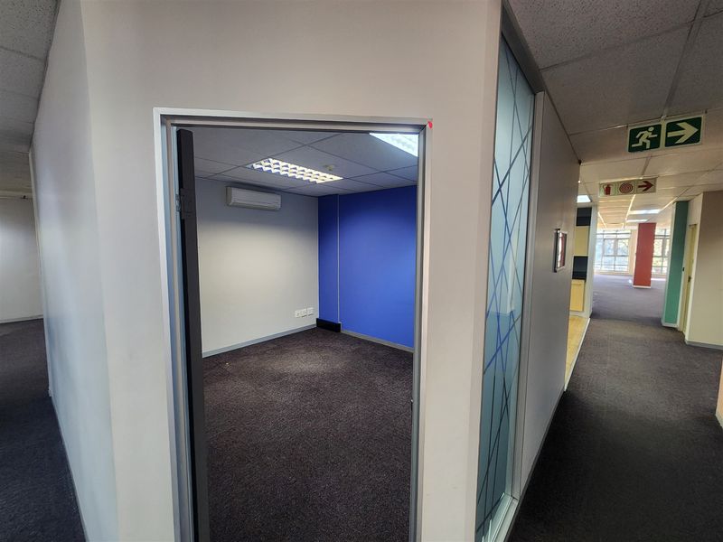A vacant office space featuring carpeted flooring, a wall-mounted air conditioning unit, and a modern frosted glass partition, situated off a main office corridor.