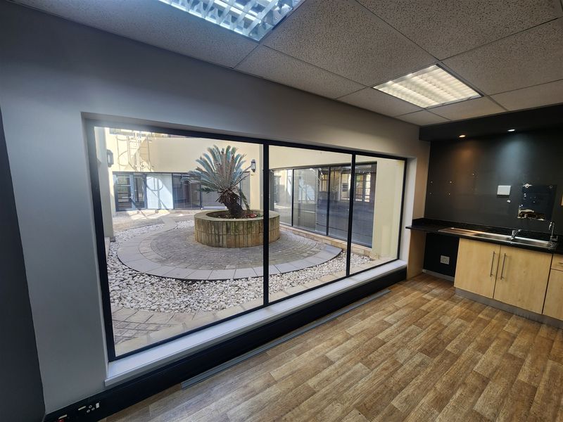 Modern office space featuring wood-look flooring, a built-in kitchenette, and a large window overlooking a central courtyard with a circular planter.