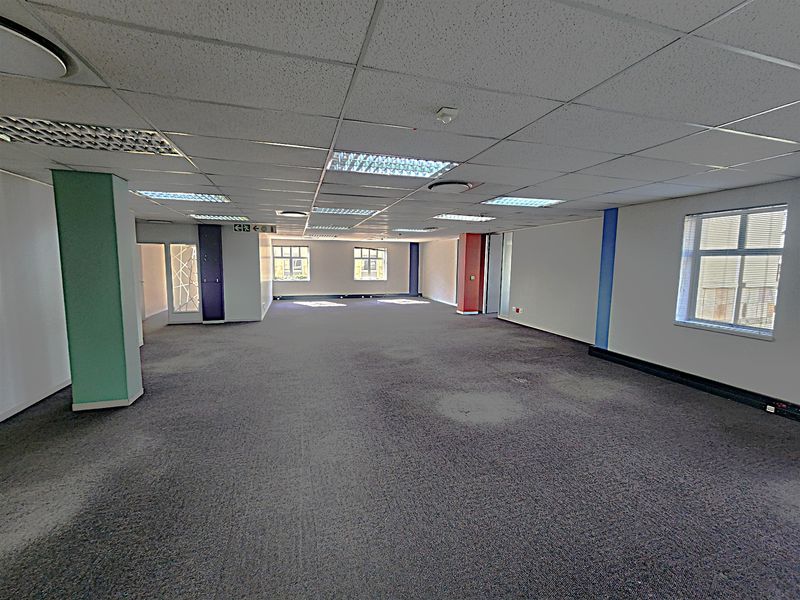 A spacious, empty open-plan office area featuring a suspended ceiling, recessed lighting, and large windows providing natural light.