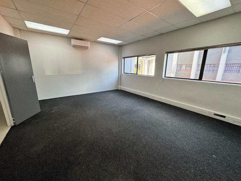 A clean, empty office space featuring dark carpeting, a suspended ceiling with recessed lighting, and a wall-mounted split-system air conditioning unit.