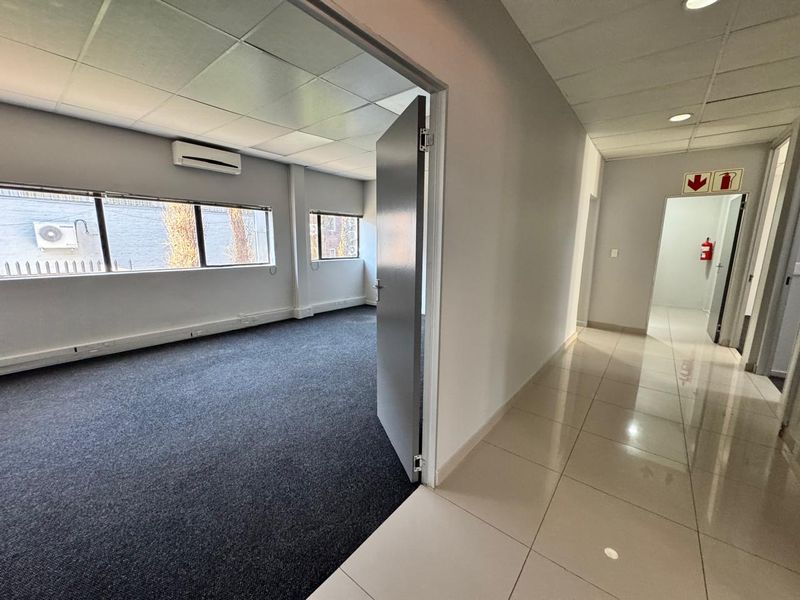 A bright, vacant office space featuring a combination of carpeted and tiled flooring, suspended ceilings, and a wall-mounted air conditioning unit.