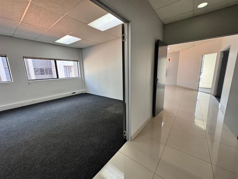 An empty office space featuring dark carpeting, a suspended ceiling with recessed lighting, and an adjacent tiled hallway.