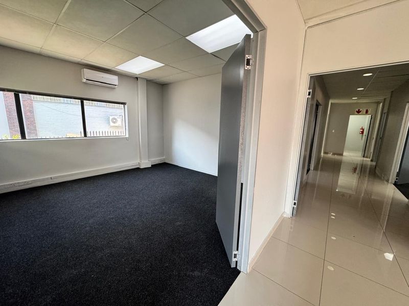 A clean, empty office space featuring dark carpet flooring, a suspended ceiling with recessed lighting, and a wall-mounted air conditioning unit.