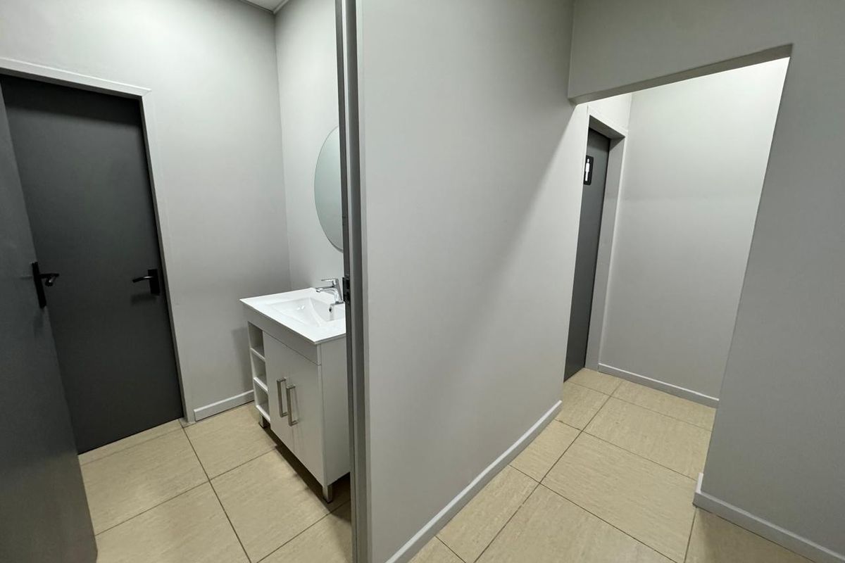 A clean, modern office bathroom area featuring a white vanity cabinet, ceramic basin, and neutral tiled flooring.