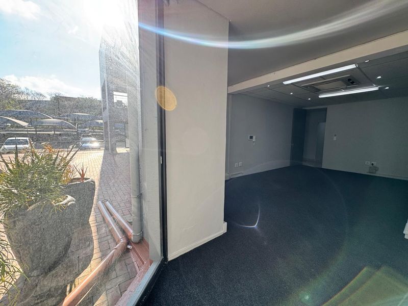 A bright, empty office space featuring carpeted flooring, recessed ceiling lighting, integrated air conditioning, and a large window providing natural light.