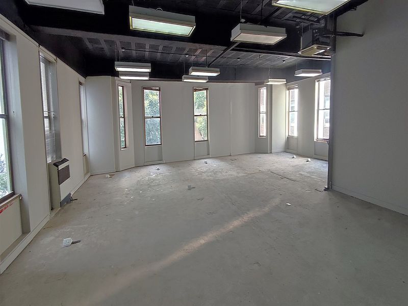 An open-plan office space featuring an exposed black ceiling, multiple windows providing natural light, and wall-mounted heating units.