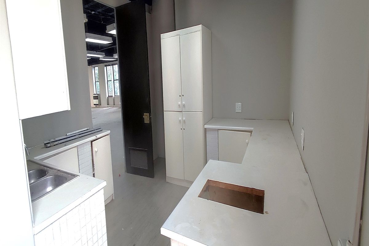 A small, empty office kitchenette featuring built-in cabinetry, laminate countertops, and a double stainless steel sink, requiring minor updates.
