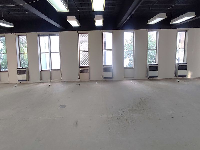 An expansive, empty office space featuring exposed ceiling beams, multiple vertical windows, wall-mounted HVAC units, and industrial-style concrete flooring.