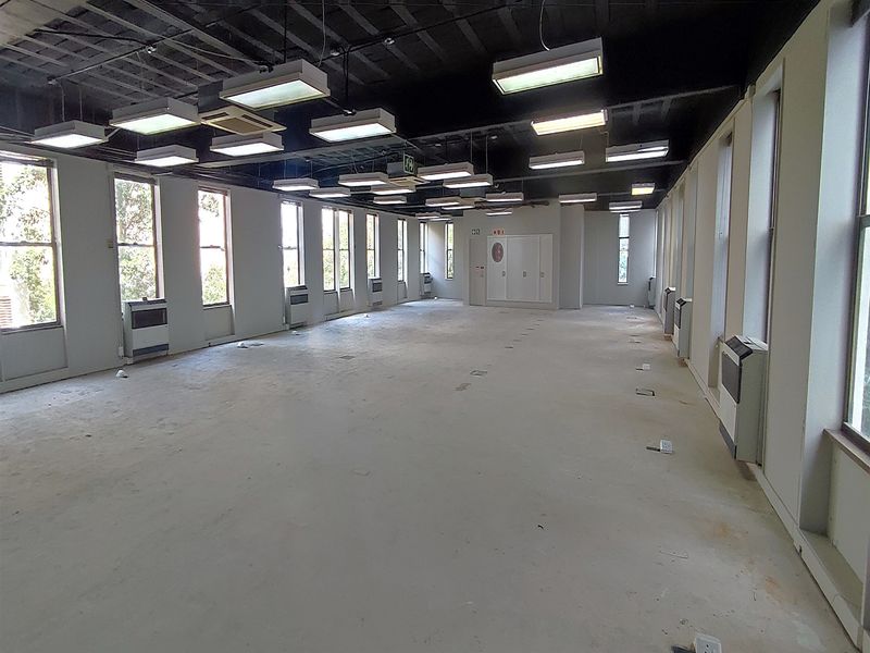 A spacious, open-plan office area featuring exposed ceilings, multiple wall-mounted air conditioning units, and large windows providing ample natural light.