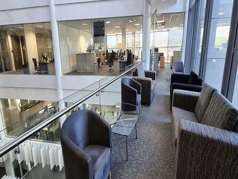 A modern office hallway featuring a glass balustrade overlooking the atrium, carpeted flooring, and floor-to-ceiling windows providing ample natural light.