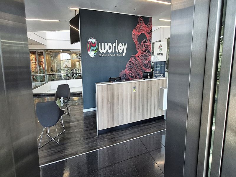 Modern office reception area featuring a branded desk, laminate flooring, and glass partitions, viewed from the elevator entrance.