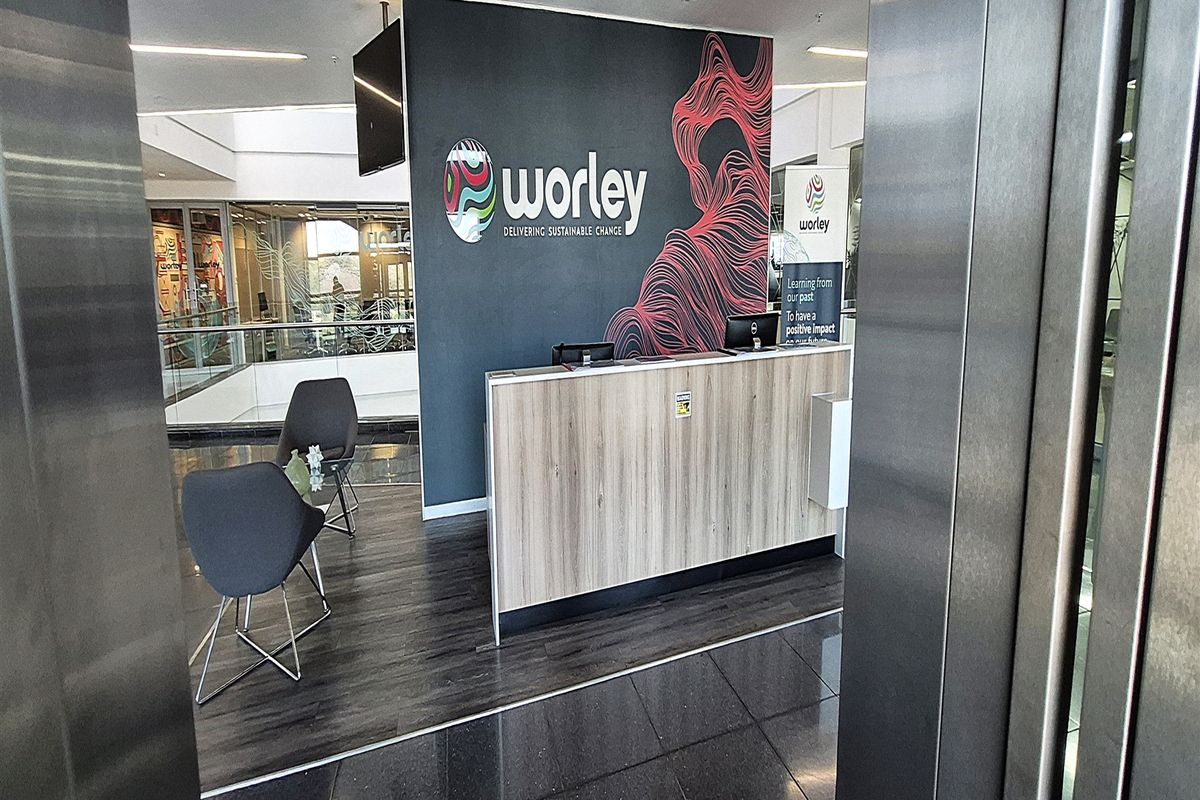Modern office reception area featuring a branded desk, laminate flooring, and glass partitions, viewed from the elevator entrance.