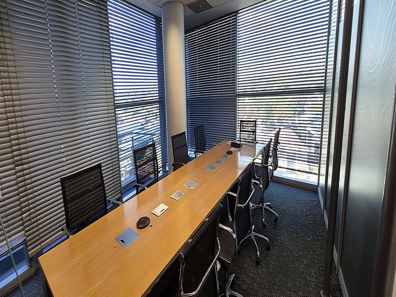 A professional office meeting room featuring large windows with venetian blinds, a central conference table, carpeted flooring, and modern glass partitioning.