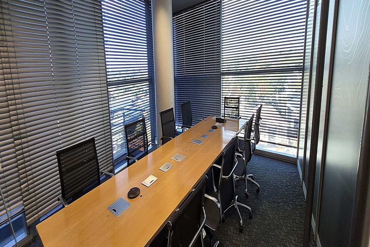 A professional office meeting room featuring large windows with venetian blinds, a central conference table, carpeted flooring, and modern glass partitioning.