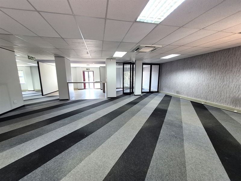 A spacious, vacant open-plan office area featuring striped carpet tiles, a suspended ceiling with recessed lighting, and glass partition doors.