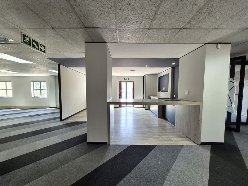 A modern, open-plan office space featuring a built-in kitchenette, suspended ceilings with recessed lighting, and a combination of carpet tiles and laminate flooring.