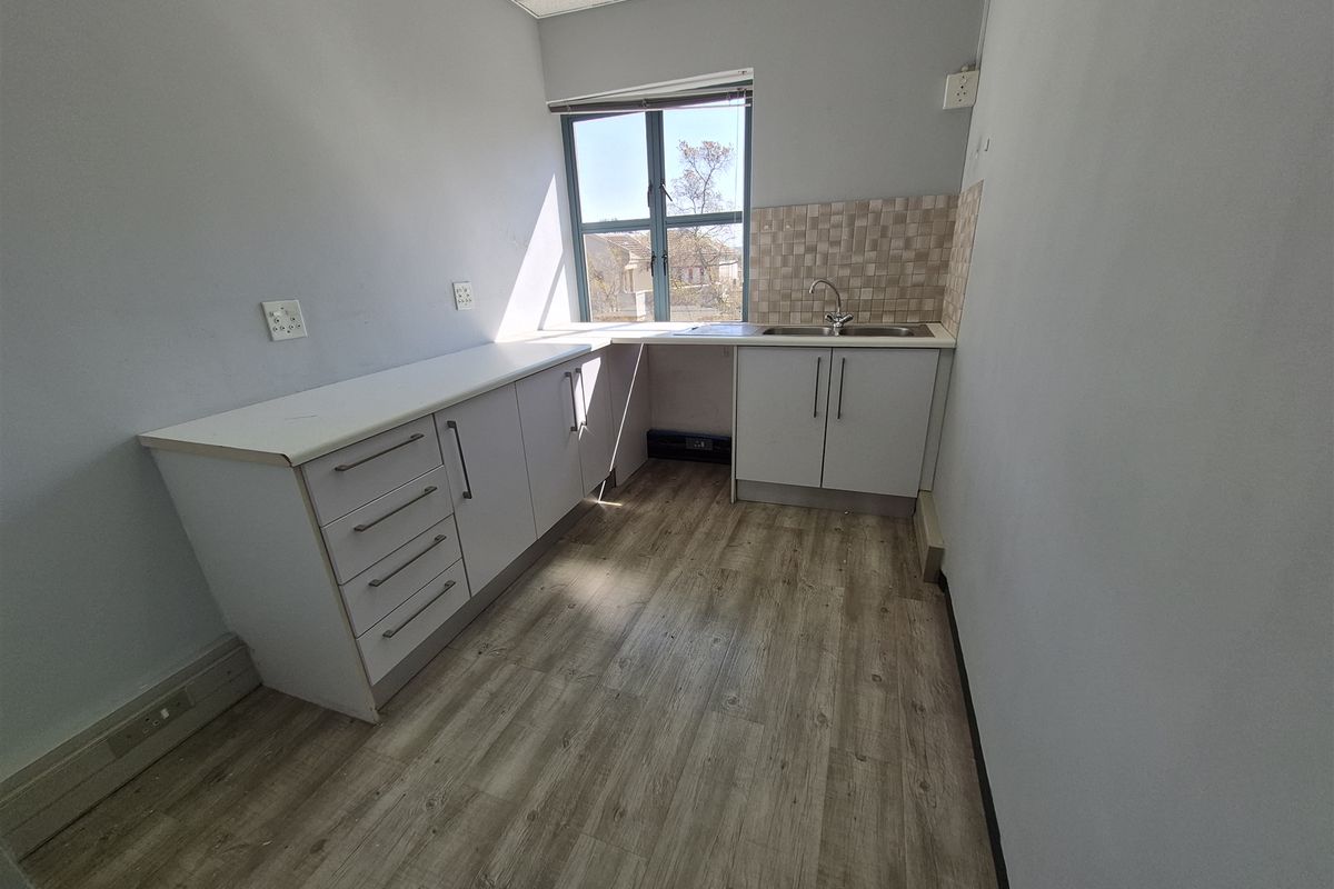 A clean, empty office kitchenette featuring white cabinetry, laminate wood-look flooring, a stainless steel sink, and a tiled backsplash under a drop ceiling.