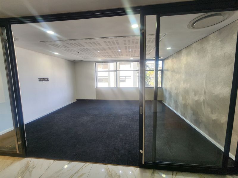 A bright, empty office space featuring a modern sliding glass partition, textured feature wall, recessed ceiling lighting, and dark carpet flooring.