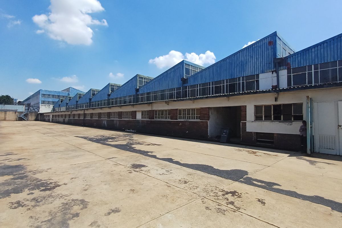 Exterior view of an industrial warehouse facility featuring a large concrete yard and sawtooth roof design with clerestory windows.