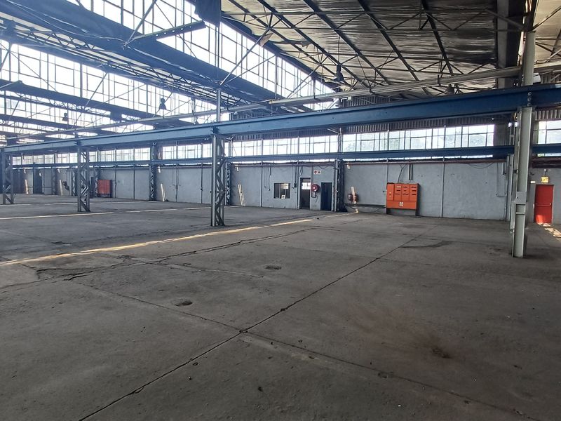 Expansive industrial warehouse space featuring high ceilings, steel structural beams, concrete flooring, and large windows providing ample natural light.