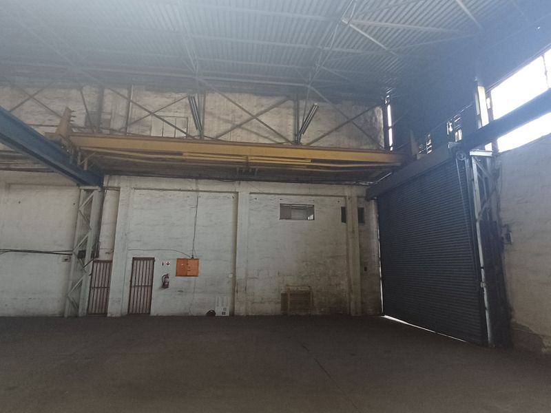 Spacious industrial warehouse featuring high ceilings, exposed steel trusses, a large roller shutter door, and an overhead crane rail system.