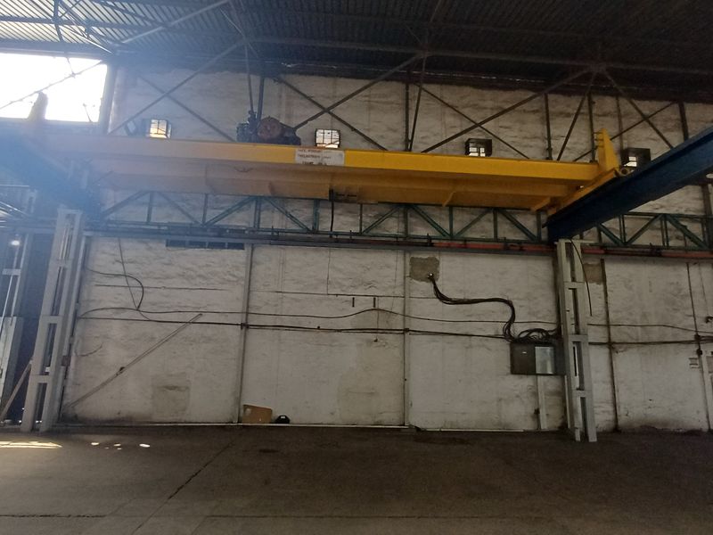 Spacious industrial warehouse interior featuring a high-capacity overhead crane system, exposed steel structural beams, and concrete flooring.