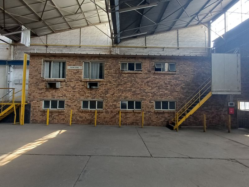 Industrial warehouse interior featuring a two-story brick office block, concrete flooring, exposed steel roof trusses, and metal staircase access.