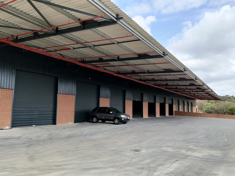 Industrial warehouse exterior featuring multiple roller shutter doors, extensive covered loading area, exposed steel roof structure, and a large concrete hardstand yard.