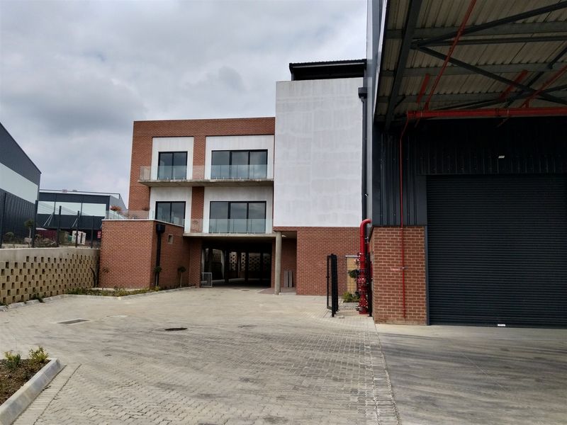 Modern industrial facility featuring a brick facade, roller shutter door, and multi-level balconies with glass balustrades, set within a paved commercial complex.