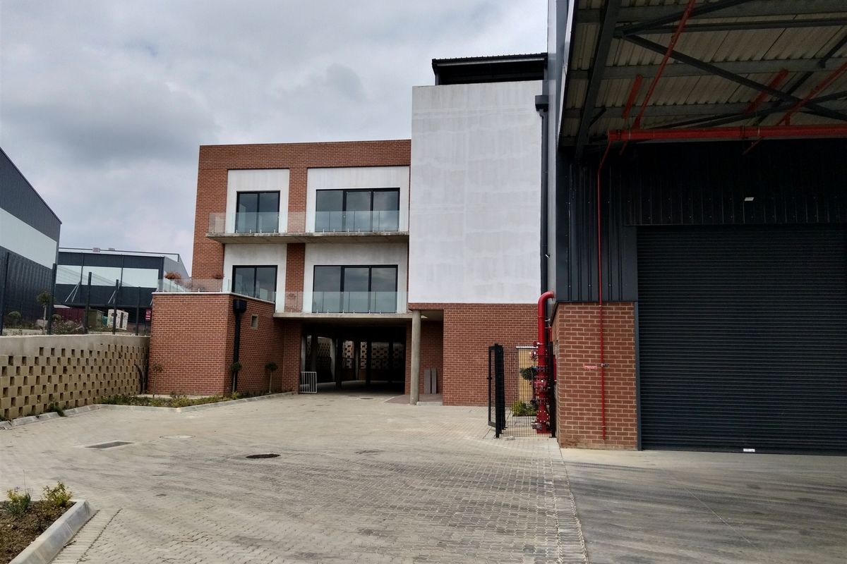 Modern industrial facility featuring a brick facade, roller shutter door, and multi-level balconies with glass balustrades, set within a paved commercial complex.