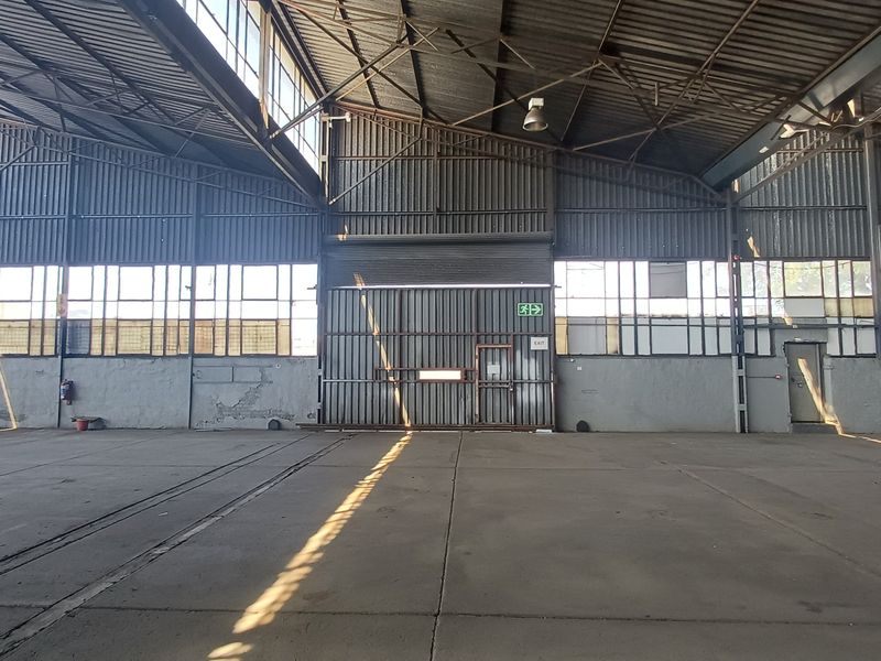 Spacious industrial warehouse interior featuring high-clearance steel truss roofing, large clerestory windows, concrete flooring, and a large sliding metal access door.