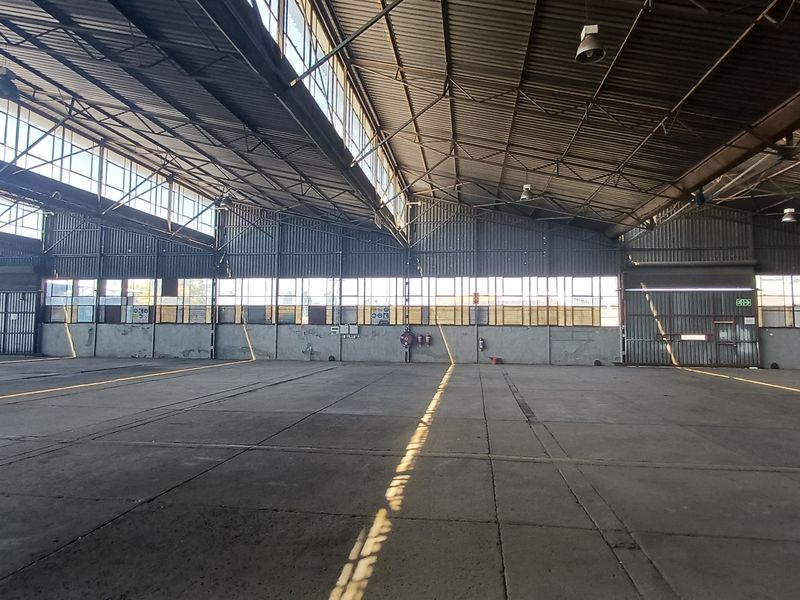 Expansive industrial warehouse space featuring high vaulted ceilings, steel truss structure, concrete flooring, and large clerestory windows providing natural light.