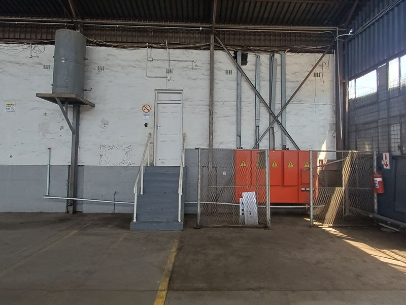 Industrial warehouse interior featuring high ceilings, concrete flooring, a raised access door with stairs, and a secured electrical distribution board enclosure.