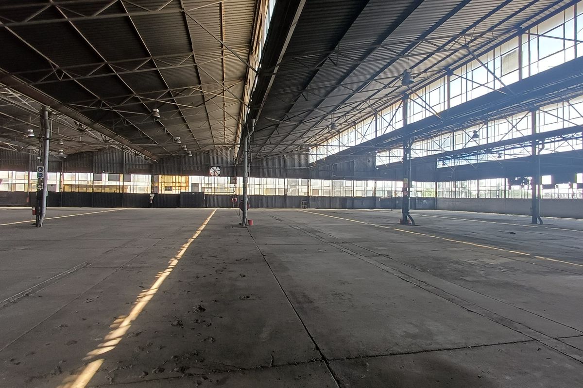 Expansive industrial warehouse space featuring high vaulted ceilings, steel structural trusses, and extensive natural light through large perimeter windows.