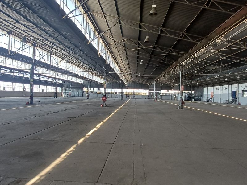 Expansive industrial warehouse space featuring high ceilings, steel truss roofing, large clerestory windows for natural light, and durable concrete flooring.